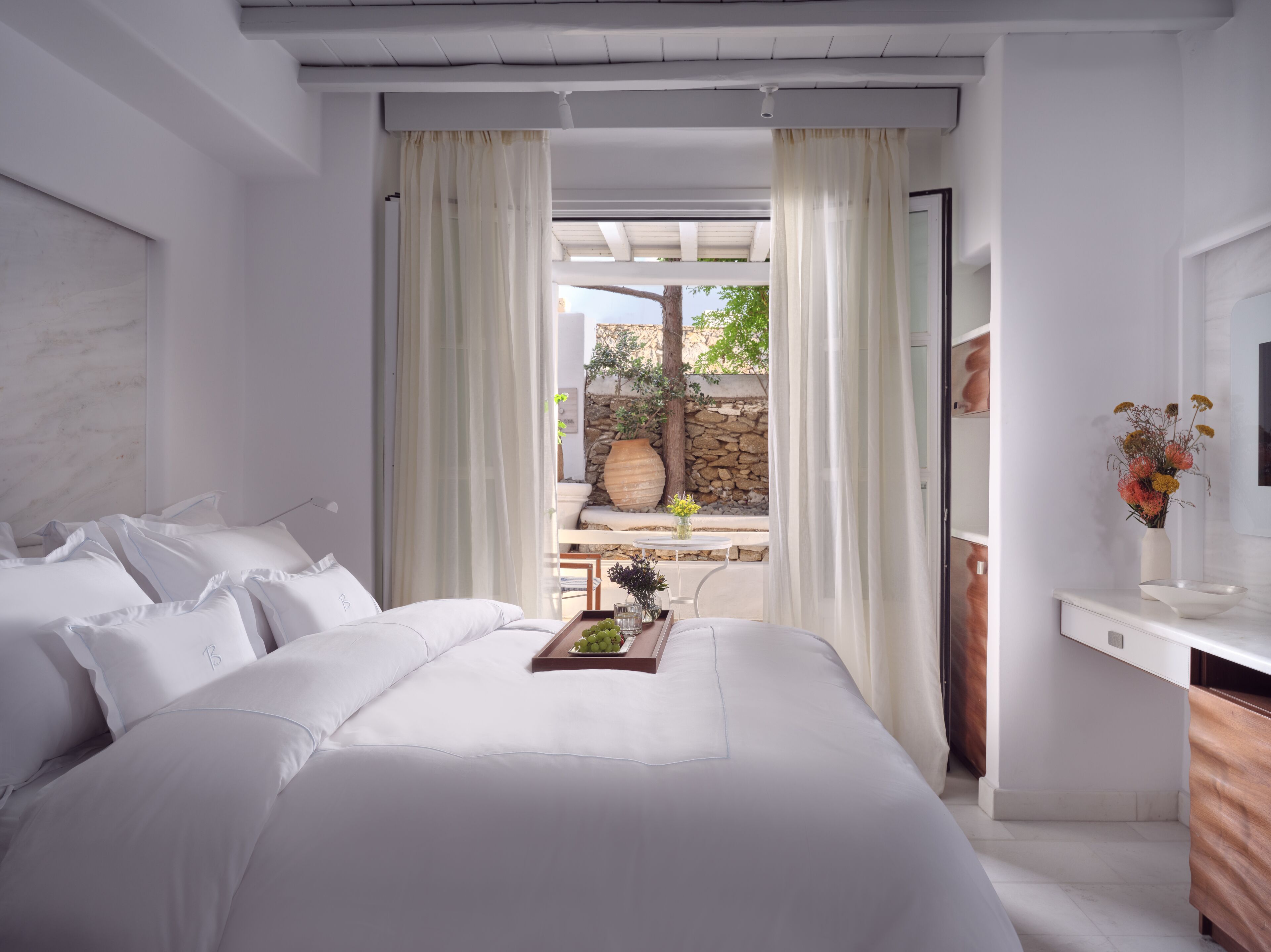 A bedroom with a large bed, a bedside table with a vase of flowers, and a view of the outdoors through a glass door.
