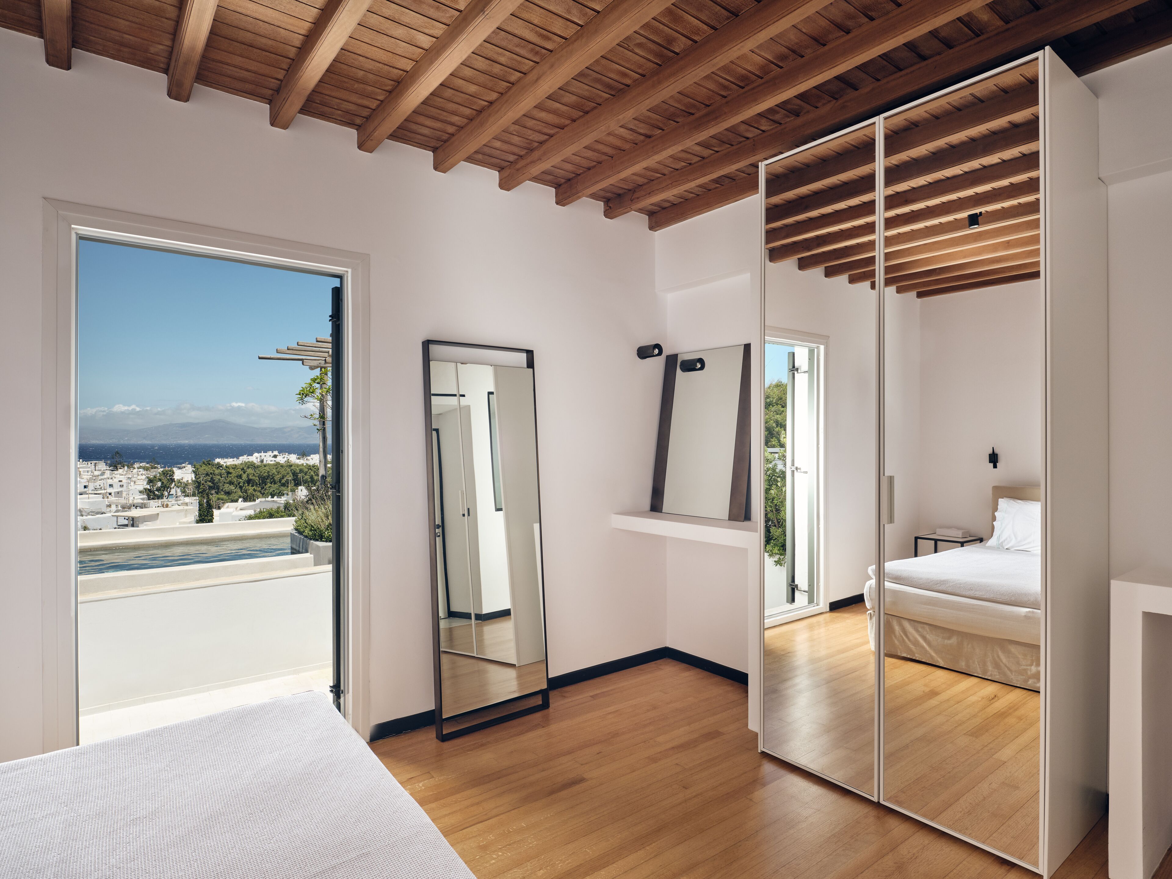 A modern bedroom with a wooden ceiling, a large mirror, a bed, and a view of a cityscape through a large window.