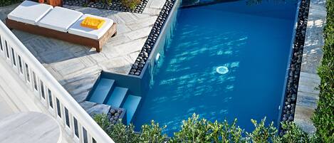 A rectangular swimming pool with a stone border, a wooden lounge chair with a white cushion, and a small wooden table with a yellow bottle.