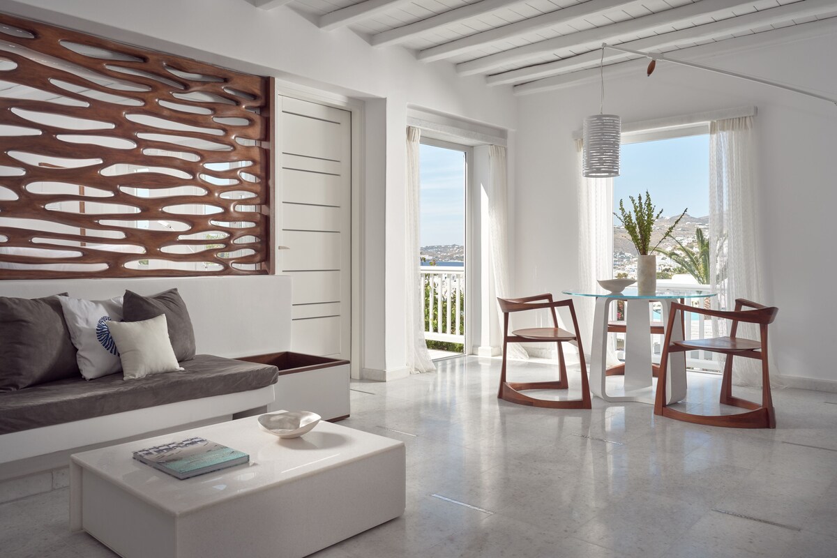 Una sala de estar moderna con un sofá blanco, una mesa de centro de vidrio y una mesa de comedor de madera con sillas. En la pared hay un panel decorativo de madera, y se ve el mar a través de ventanas grandes.