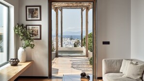 A modern living room with a wooden floor, a beige sofa, a glass coffee table, and a view of a cityscape through a wooden archway. - Belvedere Hotel - The Leading Hotels of the World  (Mykonos)