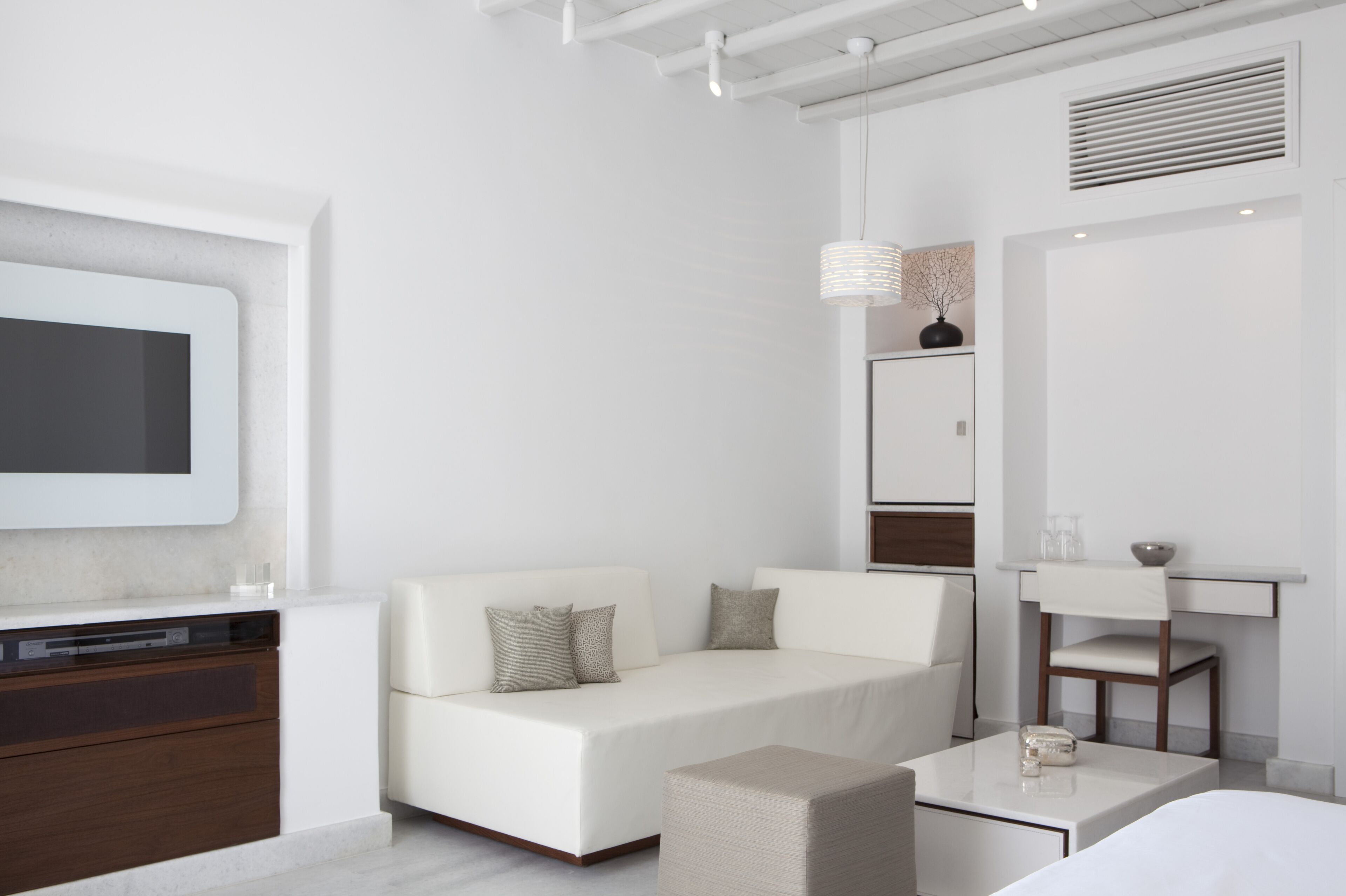 Quarto de hotel moderno com um sofá branco, uma mesinha, uma televisão e um espelho.