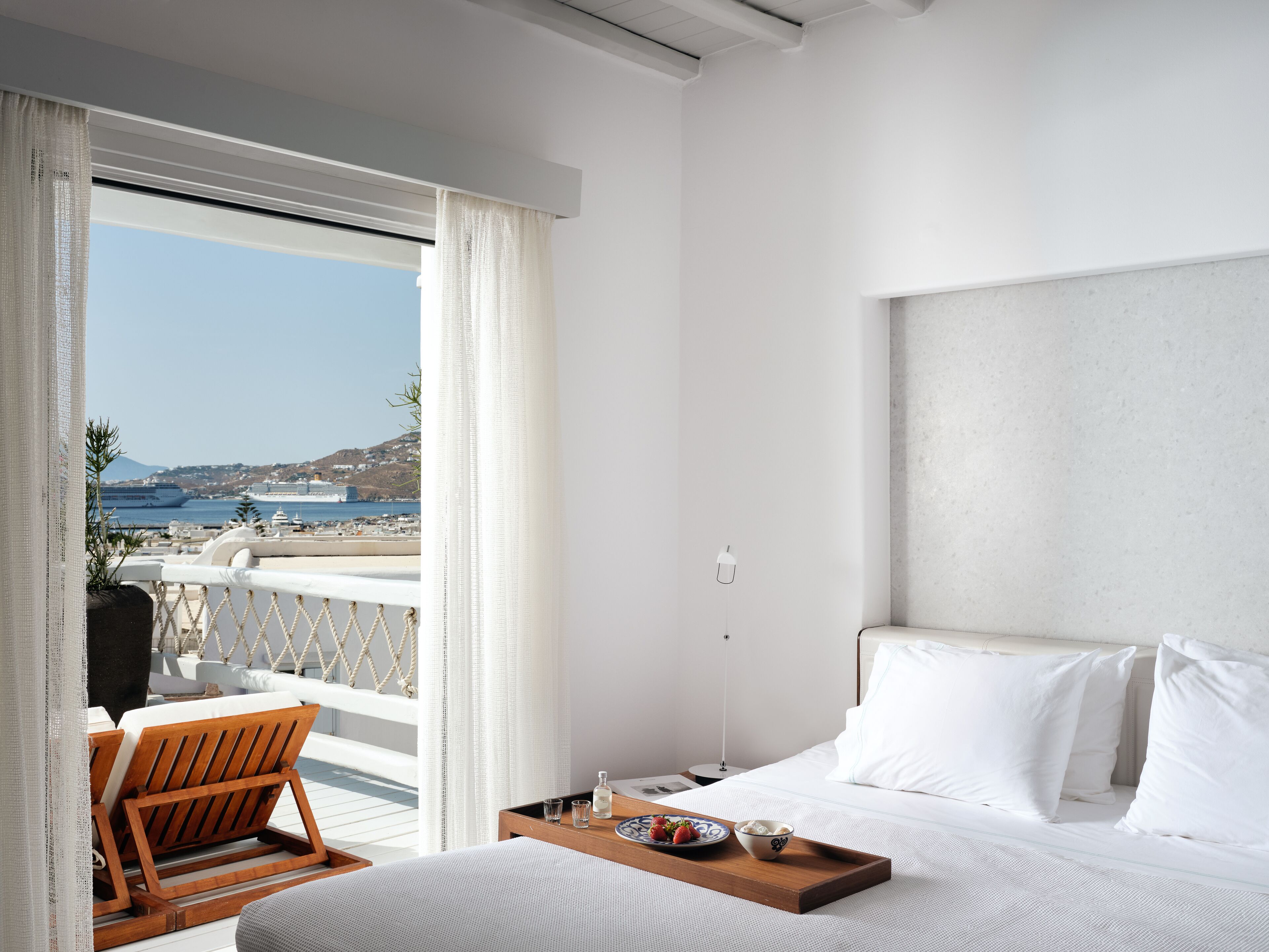 A hotel room with a bed, a wooden chair, a tray with food, and a view of the sea and ships.