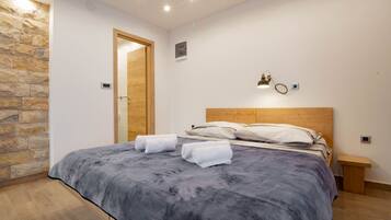 Un dormitorio con una pared de acento de piedra, un cabecero de madera, una cama con una colcha gris, dos mesitas de noche y un pequeño banco de madera.