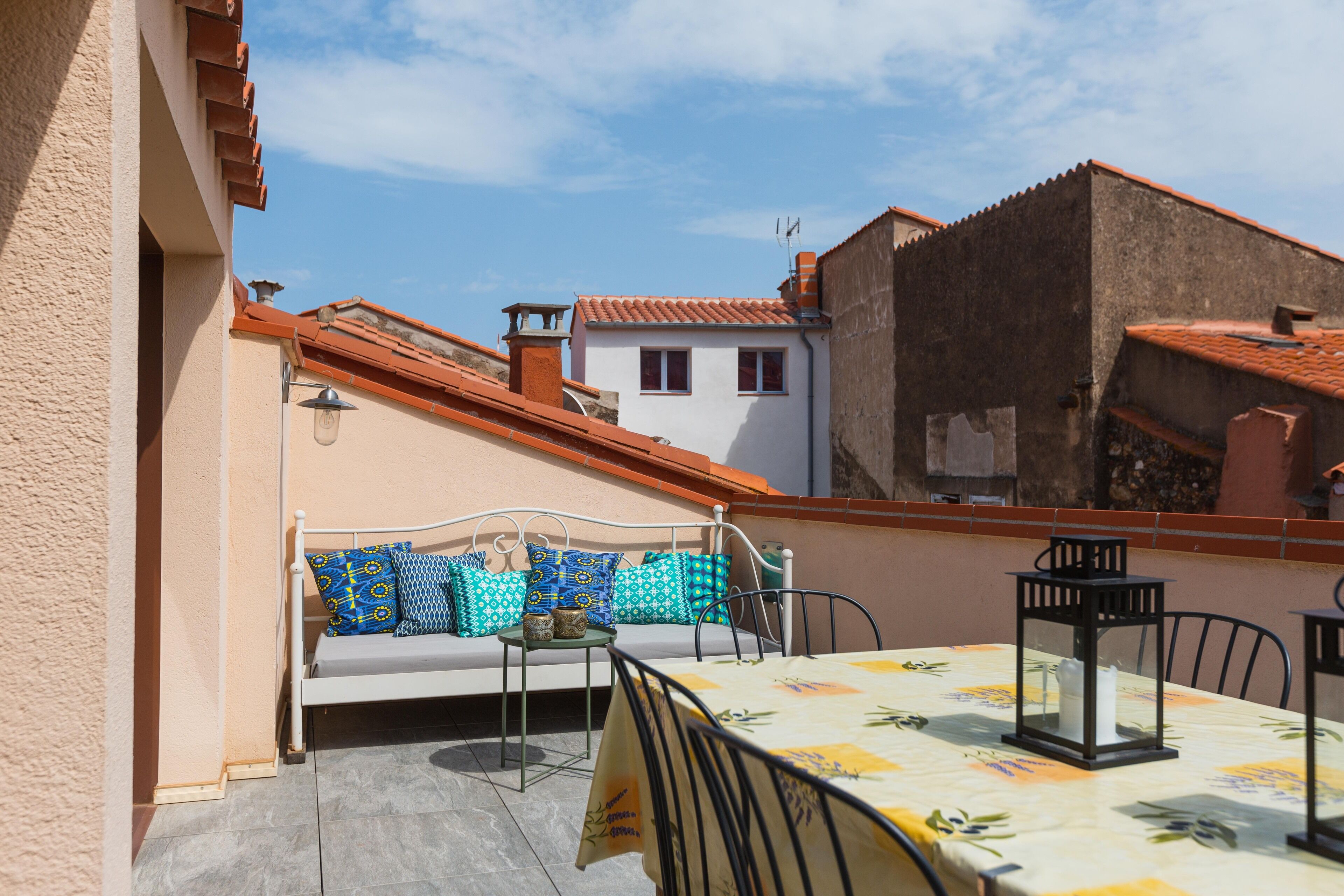 A rooftop terrace with a white sofa, blue patterned cushions, a table with a floral tablecloth, and a lantern.