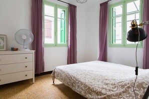 Une chambre à coucher comprenant un lit, une commode, un ventilateur et deux fenêtres avec des cadres verts et des rideaux violets.