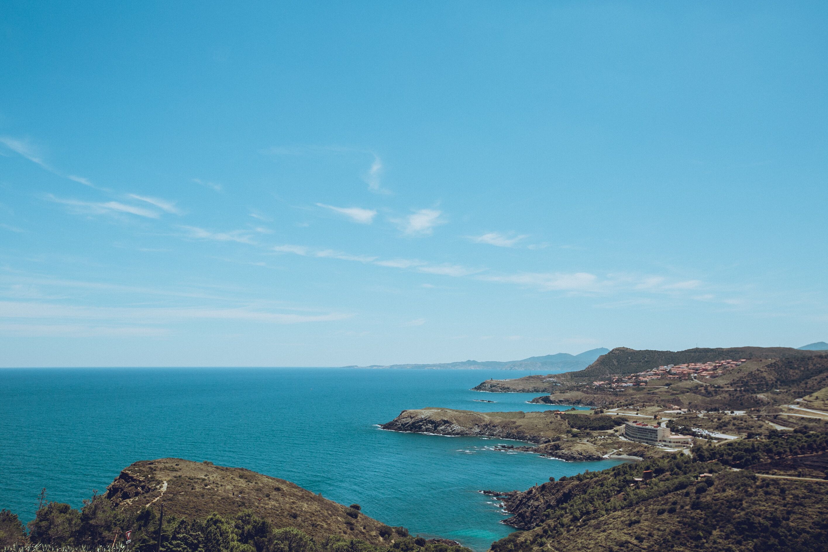 A coastal landscape with a clear sky, rocky terrain, and a distant view of a town with buildings.