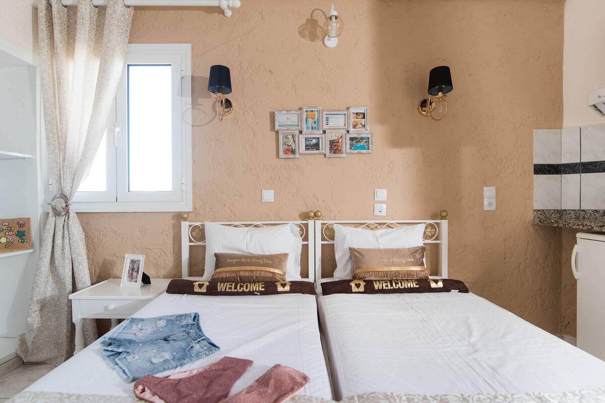 A hotel room with two beds, a small table, a window with curtains, wall-mounted lights, and a wall with framed pictures.