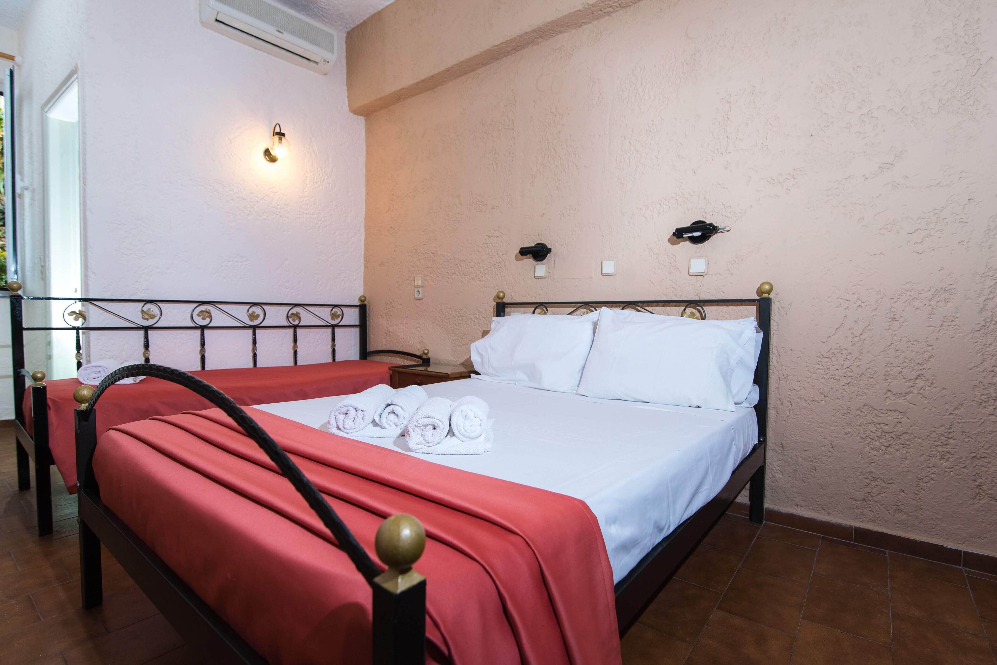 A neatly made bed with white linens and a red bedspread, a wall-mounted light fixture, and a small bedside table with a lamp.