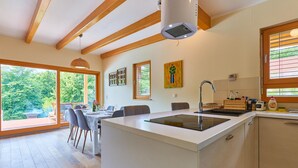 Een moderne keuken met een centraal eiland, houten balken, grote ramen en een eetgedeelte met uitzicht op de omgeving.
