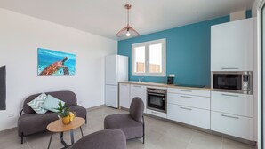 A modern kitchen with white cabinets, a microwave, and a stove. There is a dining area with a gray sofa and a small round table with a pineapple.