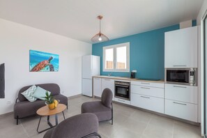 A modern kitchen with white cabinets, a microwave, and a stove. There is a dining area with a gray sofa and a small round table with a pineapple.