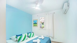 A room with two beds, a ceiling fan, an air conditioner, a window with curtains, and a framed picture on the wall.