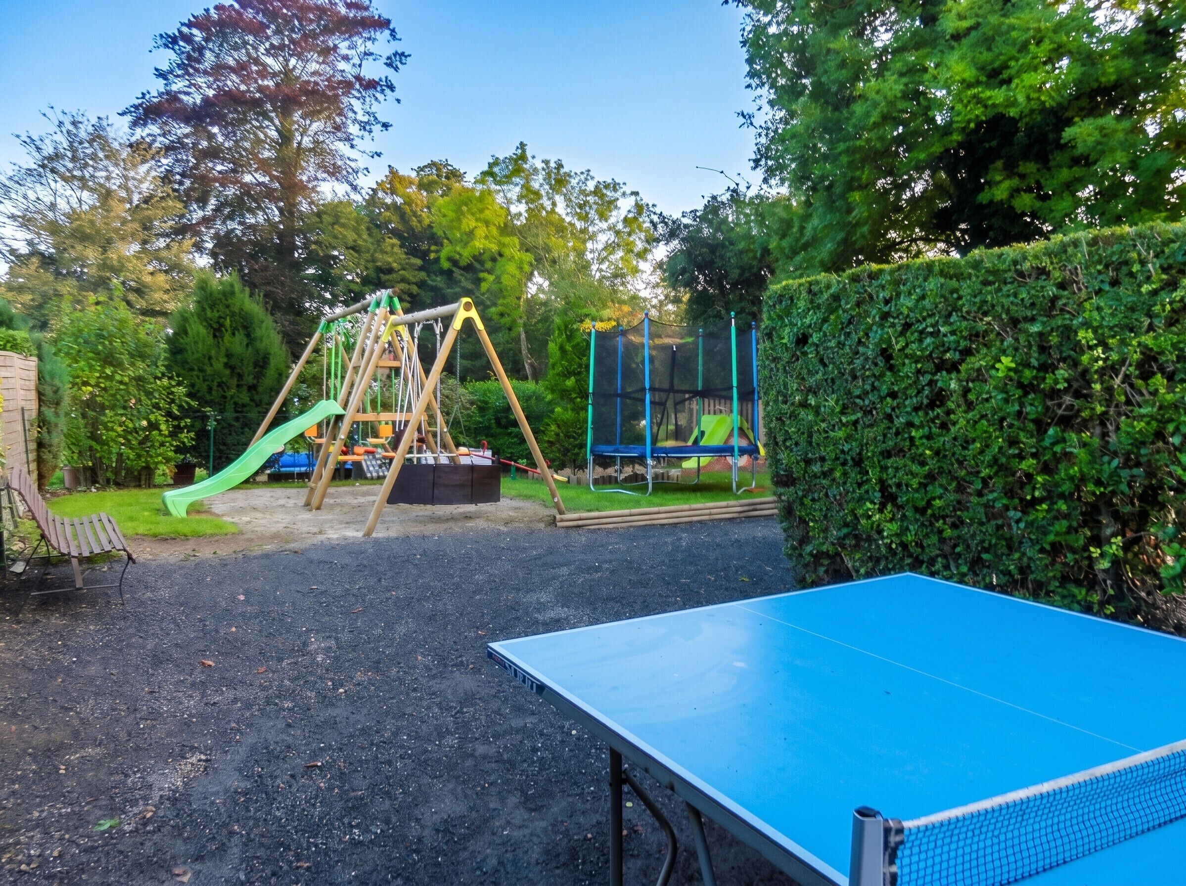A playground with swings and a slide, a tennis table, and a hedge.