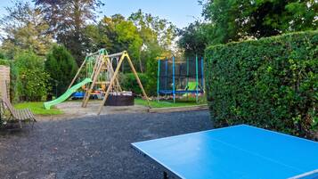 A playground with swings and a slide, a tennis table, and a hedge.