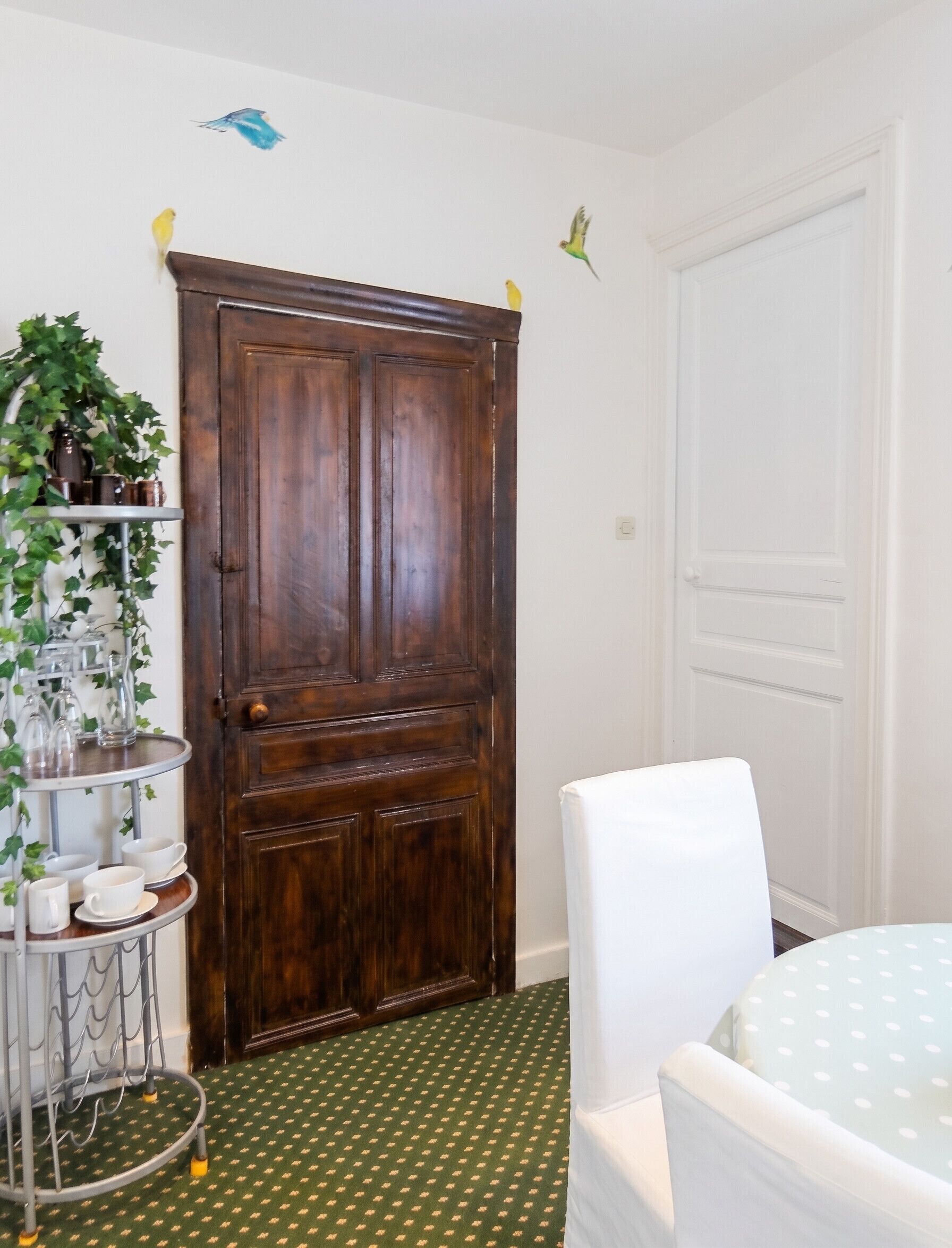 A room with a wooden wardrobe, a white chair, a round table with a polka dot tablecloth, and a metal shelf with glassware and a plant.