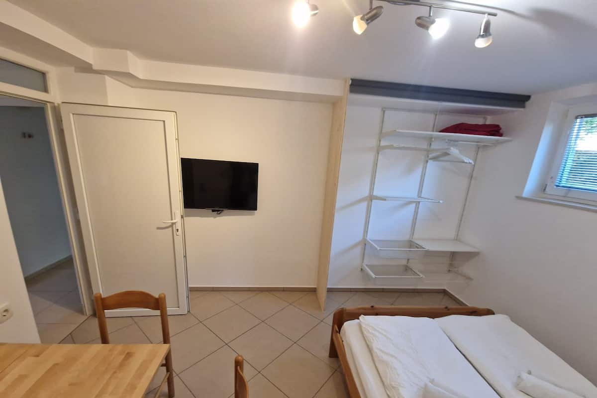 Ein kleiner, gut beleuchteter Raum mit einem Bett, einem Schreibtisch, einem Stuhl, einem an der Wand befestigten Fernseher und einer Regalanlage.