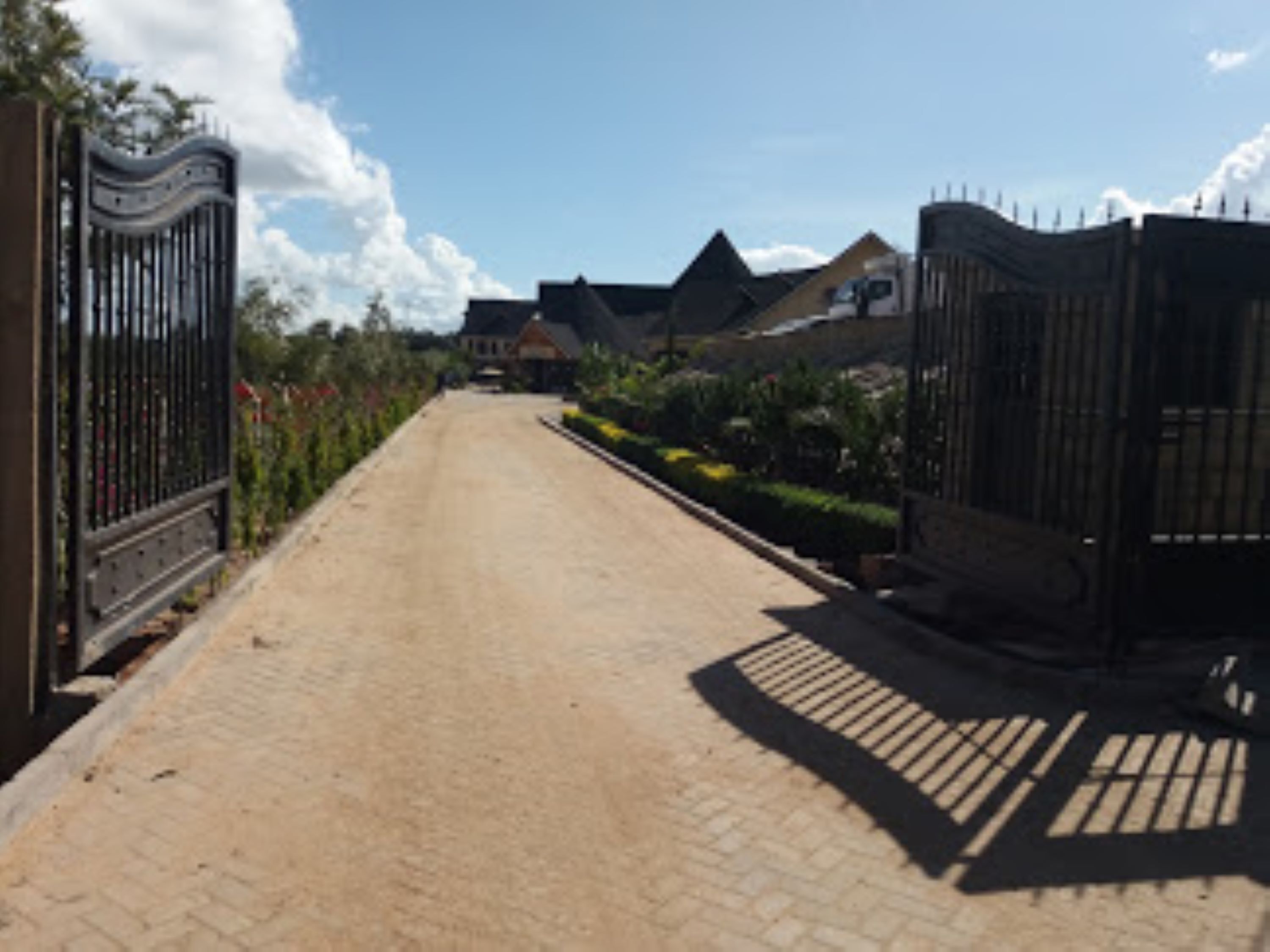 Une allée pavée mène à une entrée fermée par un portail, avec une maison visible au loin.