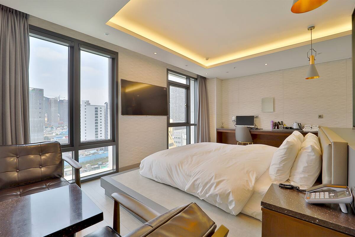 A modern hotel room with a large bed, a desk, and a view of the cityscape through large windows.