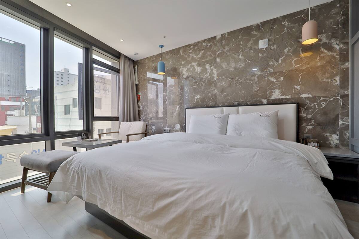 A modern hotel room with a large bed, a bedside table, a chair, and a view of the city through large windows.
