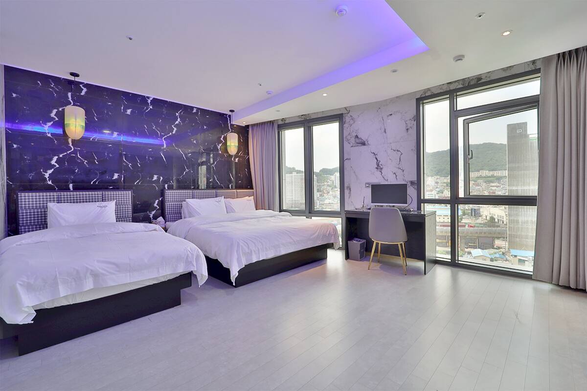 A modern hotel room with two beds, a desk, and large windows offering a city view.