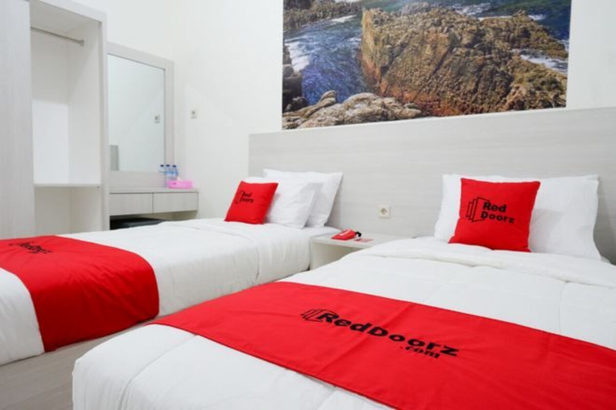 A hotel room with two beds, each with a red and white bedspread and pillows, a white wardrobe, a nightstand, and a painting of a rocky coastline on the wall.