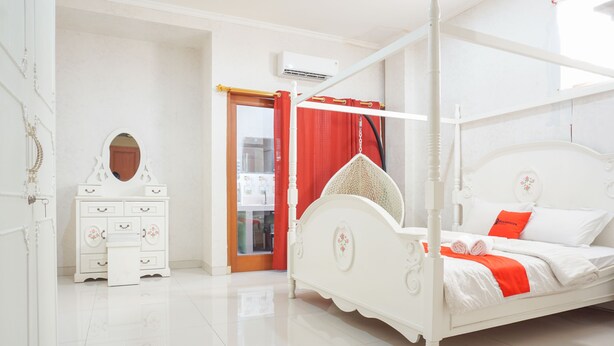Een wit hemelbed, een witte dressoir met lades, een spiegel en een rood gordijn.