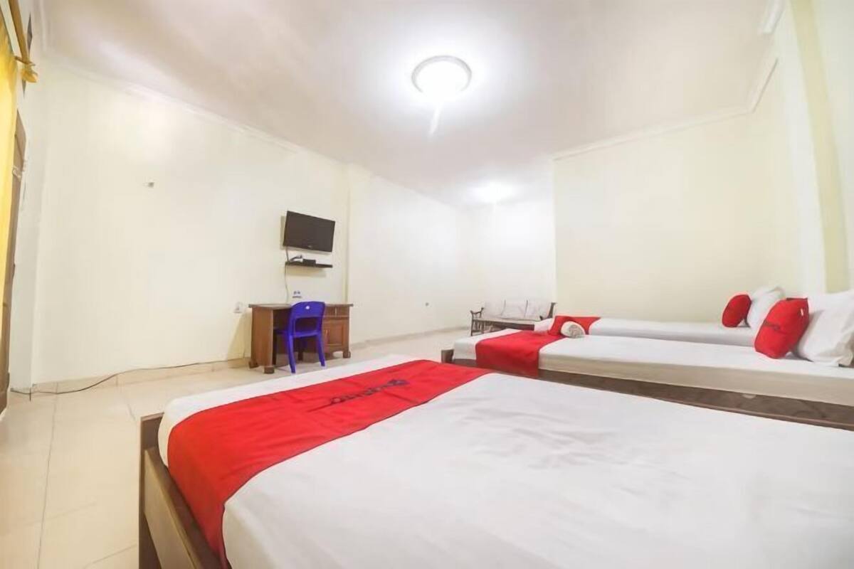 A hotel room with three beds, a desk with a chair, a television, and a red and white bedspread.