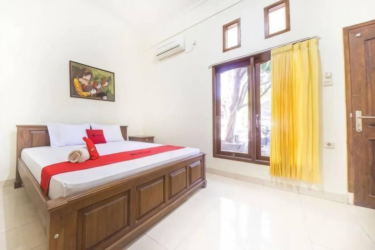 A bedroom with a wooden bed, a window with yellow curtains, a door, and a wall-mounted air conditioner.