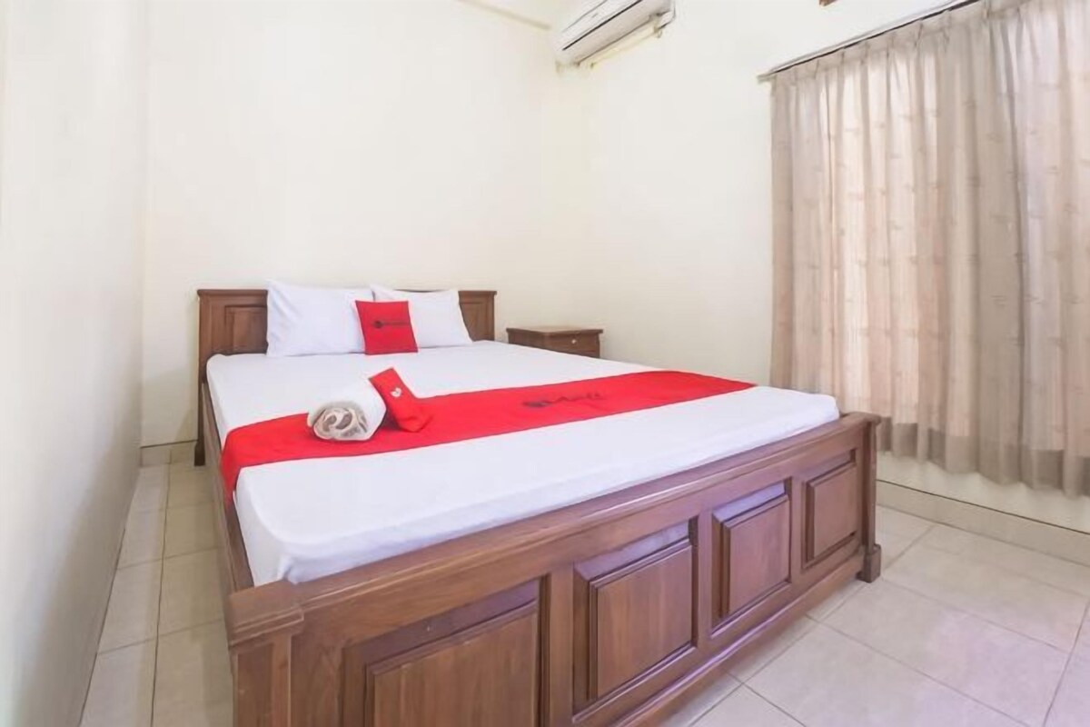 A neatly made bed with a red accent, a wooden headboard, and a window with curtains.