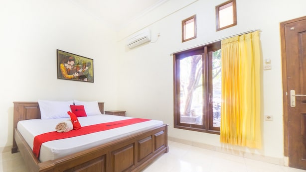 A neatly made bed with a red and white bedspread, a wooden headboard, and a single red pillow. There is a framed picture on the wall above the bed, and a window with yellow curtains to the right.