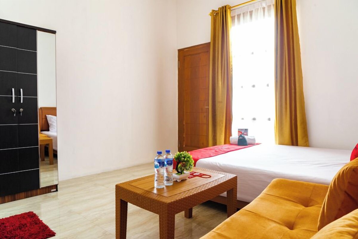 A hotel room with a bed, a wooden wardrobe, a small table with water bottles, a red rug, and a yellow armchair.