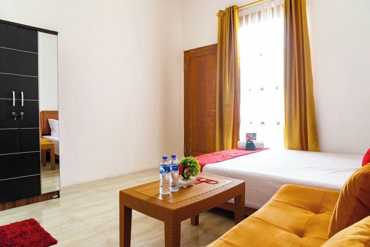 A hotel room with a bed, a wooden wardrobe, a small table with water bottles, a red rug, and a yellow armchair.