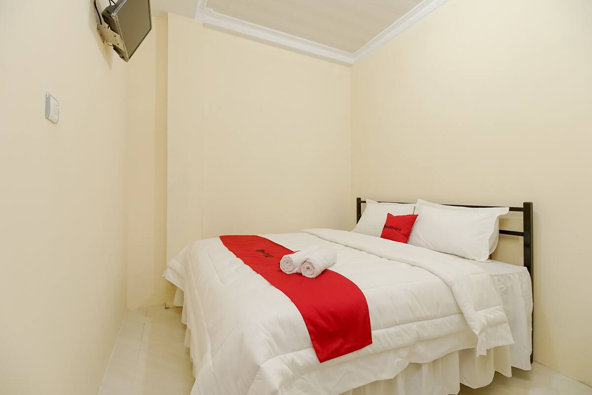 A neatly made bed with white bedding and a red accent, a wall-mounted light fixture, and a plain wall.