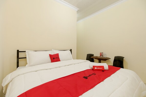 A neatly made bed with a white and red bedspread, a red pillow, and a small table with a bottle and a red towel. - RedDoorz near Tahura Berastagi (Berastagi)