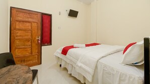 Een kamer met een houten deur, een bed met wit beddengoed en rode accenten, een klein tafeltje en een televisie aan de muur.
