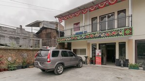 Een gebouw met twee verdiepingen, voorzien van een balkon versierd met rood-witte patronen, een geparkeerde auto en potplanten.