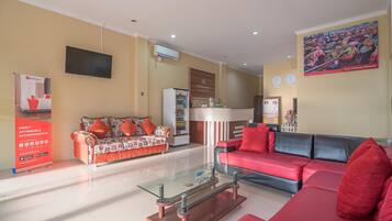 A hotel lobby with a reception desk, a television mounted on the wall, a refrigerator, and two sofas with red cushions.