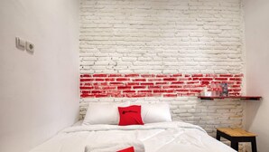 Habitación de hotel con pared de ladrillo blanco, cama con ropa de cama blanca y almohadas rojas, mesita de noche de madera y una pequeña mesa con una bandeja roja.