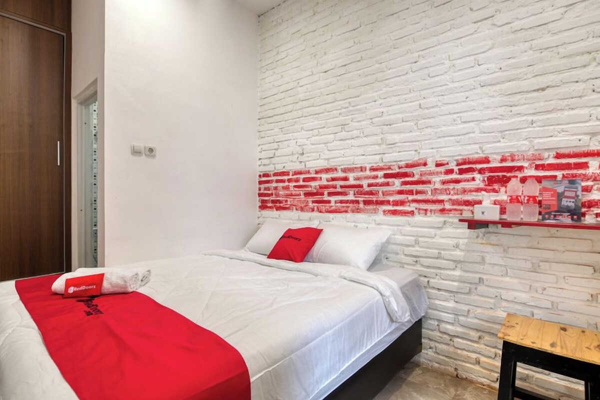 A hotel room with a white brick wall, a bed with white bedding and red accents, a wooden bench, and a small shelf with toiletries.
