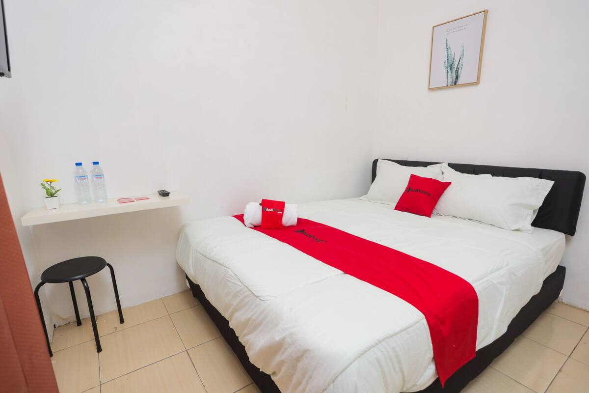 Un lit bien fait, recouvert d’une couverture blanche et rouge, un petit bureau avec une chaise et un tableau encadré accroché au mur.