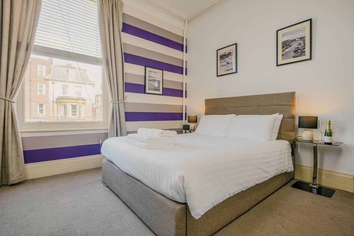 A hotel room with a bed, a nightstand, a window with curtains, and a wall with striped wallpaper and framed pictures.