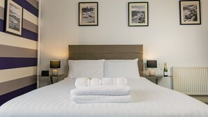 A neatly made bed with white linens and towels, a headboard, bedside tables with lamps, and framed pictures on the wall. - Rowntree Lodge (Scarborough)