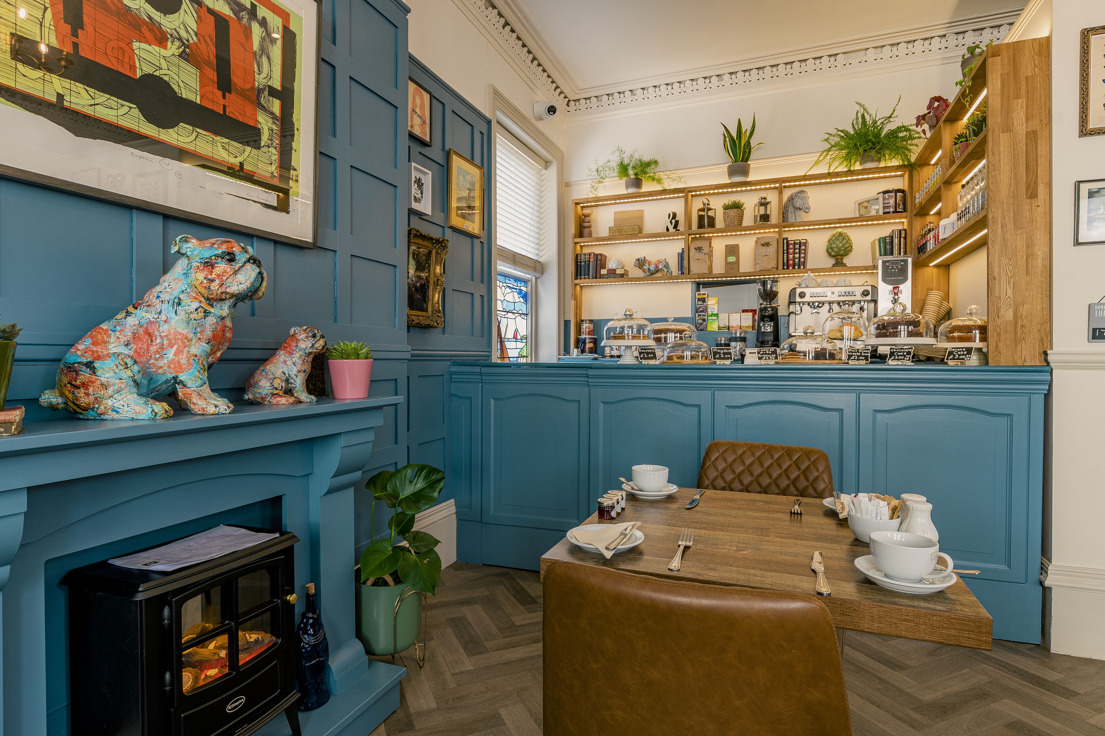 Un comedor con una mesa de madera preparada para dos personas, una pared azul con detalles decorativos y una zona de cocina con estantes y electrodomésticos.