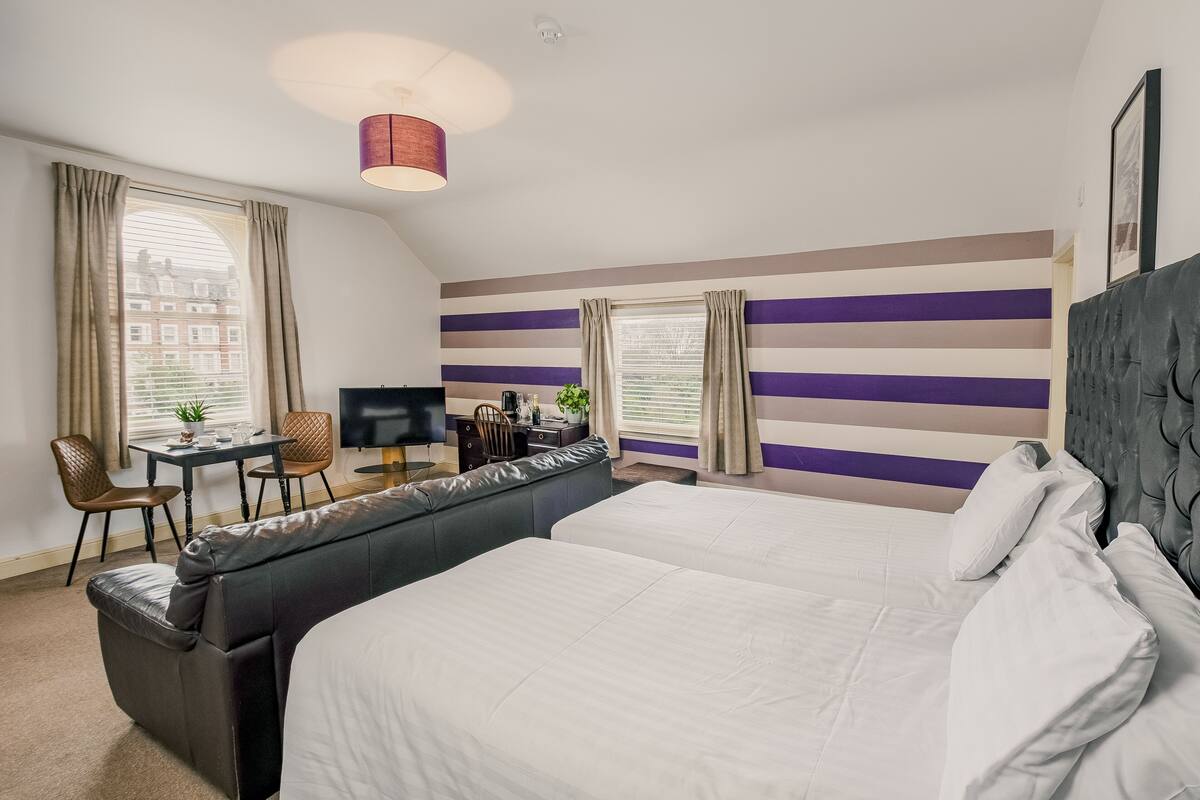 A hotel room with a large bed, a sofa bed, a desk with a chair, a television, and a window with curtains.