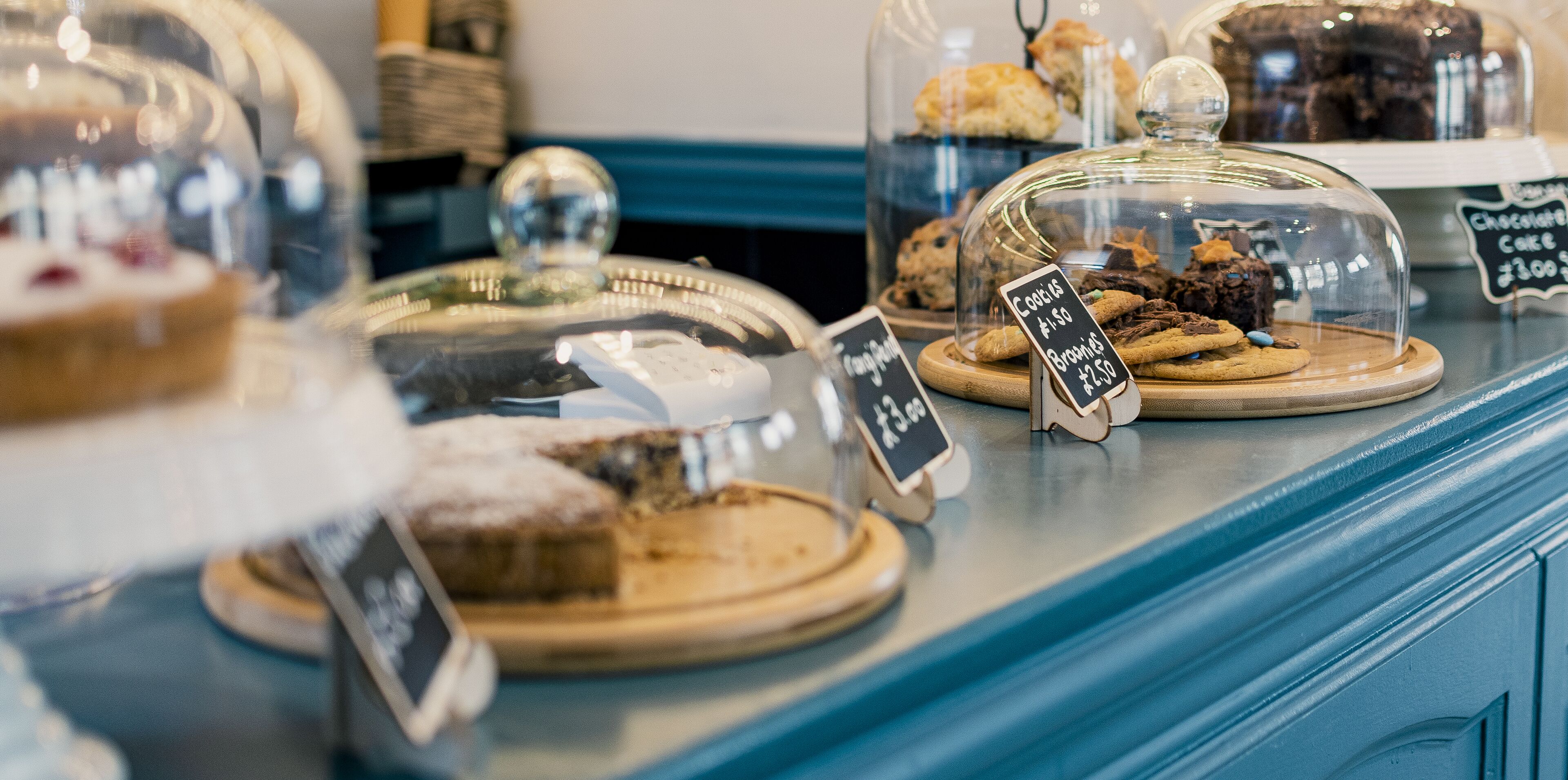 A bakery display with various baked goods under glass domes, including cakes and cookies, with price tags visible.