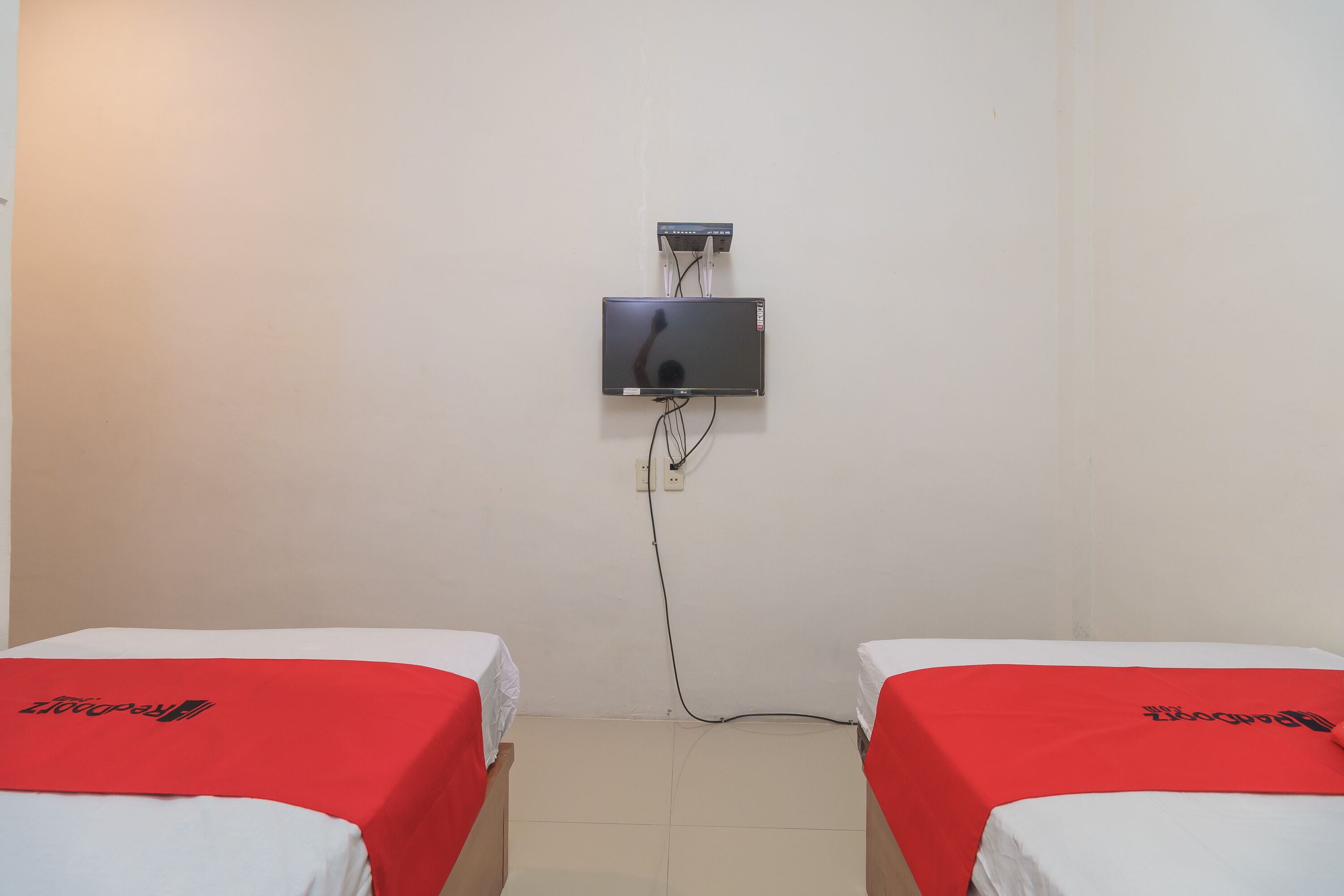 A room with two beds, each with a red blanket and a logo, a flat-screen TV mounted on the wall, and a power outlet.