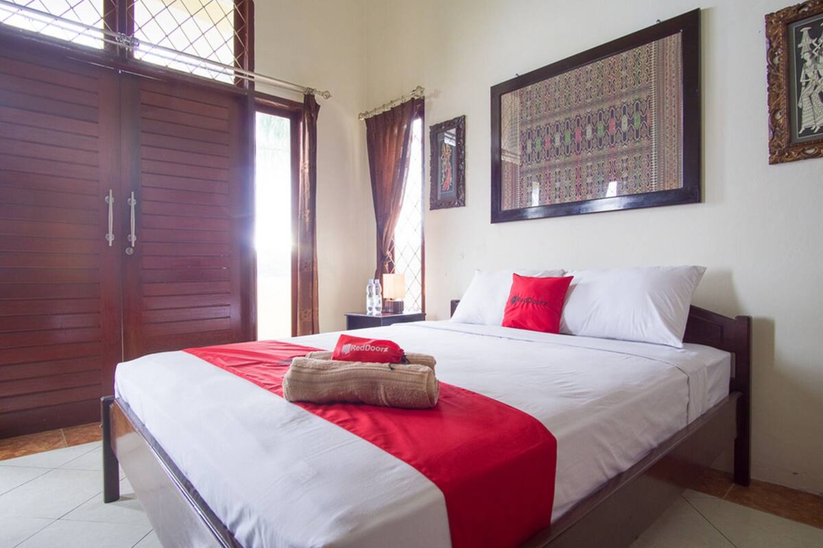 A hotel room with a large bed, a wooden wardrobe, a nightstand with a lamp, and a framed tapestry on the wall.
