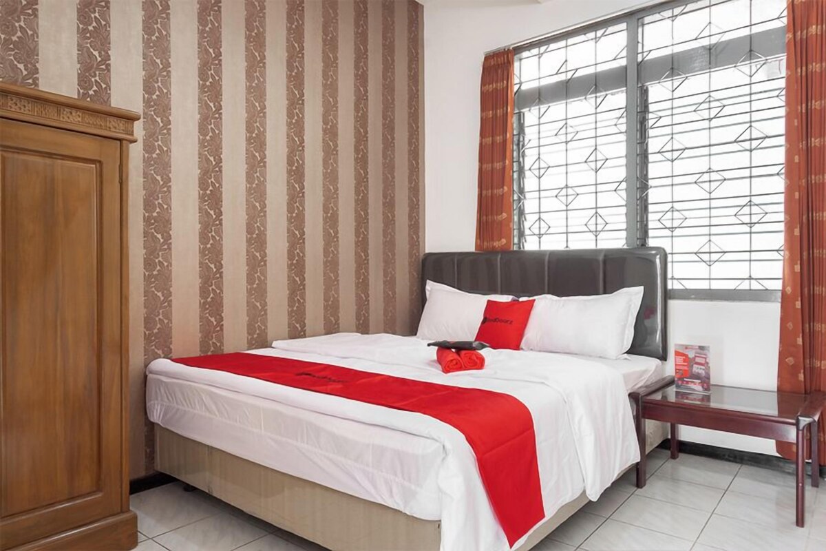 Een netjes opgemaakt bed met een rode accentkleur, een houten garderobe en een raam met behang.