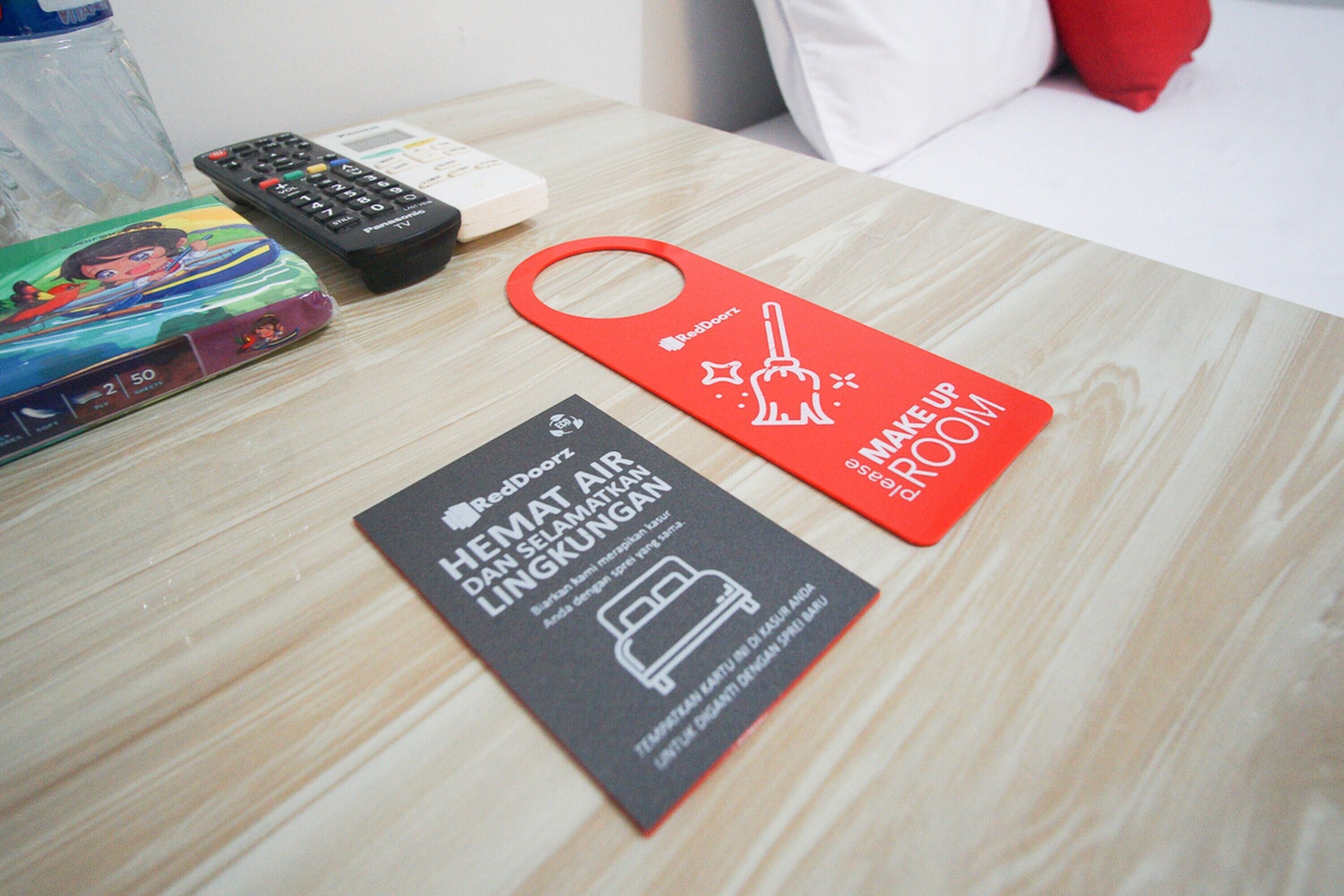A hotel room with a wooden table, a remote control, a RedDoorz sign, and a RedDoorz bag on the table.